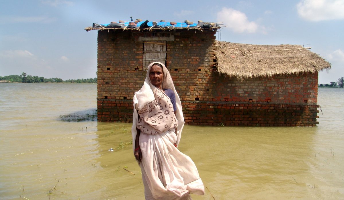 Jinsi Devi in front of her mud house that is surrounded by water with the tarpaulin on the roof which she received from CARE as part of a relief kit.